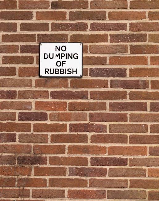 A rectangular white metal sign mounted on a red brick exterior wall, displaying the message 'NO DUMPING OF RUBBISH' in black uppercase letters, with the word 'DUMPING' partially obscured or damaged. T