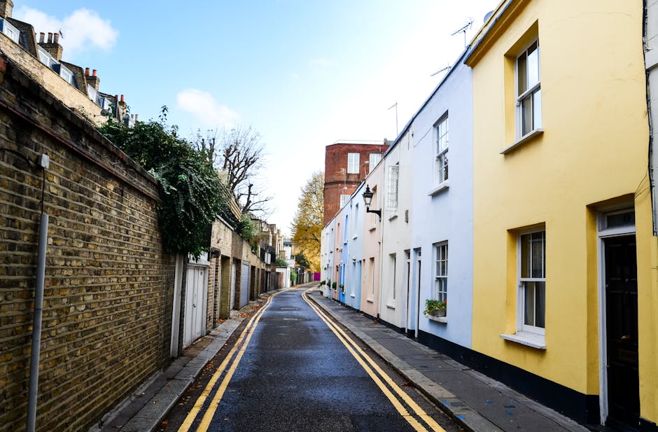 A narrow street in Forest Hill with parked cars and charming terraced houses on both sides, featuring a mix of brick and painted facades in yellow, white, and pastel colors. The street has a slight cu