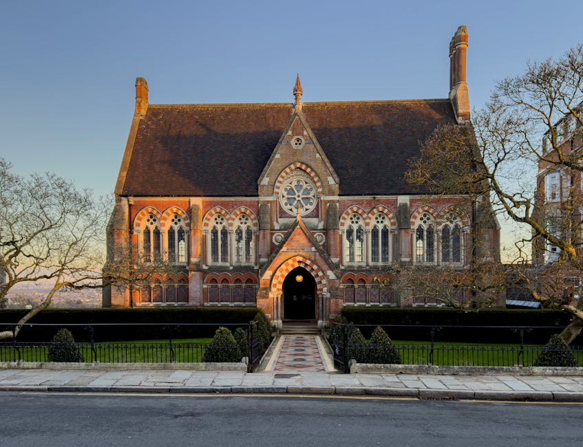 The image depicts a historic church building with Gothic architectural features, including pointed arched windows and a large circular stained glass window at the center of the facade. The building is constructed from red and gray brickwork, with a steeply pitched roof covered in red tiles. The entrance is situated at the front, featuring a rounded arch doorway framed by decorative brickwork, with a short paved pathway leading up to it. Flanking the church are leafless trees, suggesting a late autumn or winter season. The church is positioned on a paved street, with a black metal fence enclosing a small, well-maintained garden area in front of it. The scene is captured during daylight with clear blue skies overhead. The photograph is relevant to house removals and moving services by illustrating a typical landmark setting in Forest Hill, where professional furniture transport and packing services by [COMPANY_NAME] could be required for relocating or moving within or nearby this area.