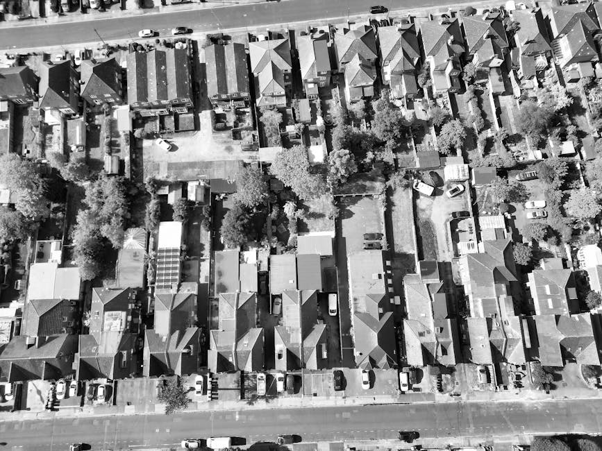 An aerial black-and-white photograph showing a residential street with closely packed houses featuring pitched roofs, small front gardens, and driveways. Vehicles, including a white van used for home relocation, are parked along the street and in driveways. The van is positioned on the pavement adjacent to the property, with some of its rear doors open, revealing a loading process involving furniture items covered with blankets and plastic wraps. Several boxes, some stacked on trolleys, and pieces of furniture such as sofas and wardrobes are visible being moved into or out of the van. Trolley carts and straps are used to facilitate the safe transport of household items. The surrounding environment includes trees, narrow pathways, and fences separating neighboring properties. This scene exemplifies the logistics involved in furniture transport and packing during house removals, with [COMPANY_NAME] providing professional removal services supporting house moves and property relocation in Forest Hill.