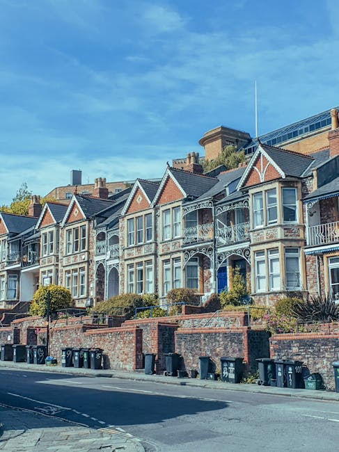 A row of Victorian-style terraced houses in Forest Hill, showing detailed facades with bay windows, decorative balconies, and brickwork. The houses are elevated on a small hill, with front gardens bordered by low brick walls and a sidewalk alongside the street. Several black wheelie bins are lined up at the base of the wall, and the sky above is clear with some light clouds. This residential area reflects typical architecture and street scenery where home relocation services by Man with Van Forest Hill might operate, supporting furniture transport and moving logistics in the local community.
