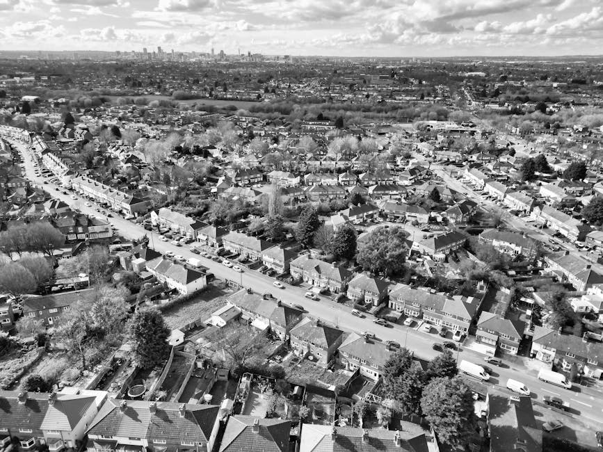 A black-and-white aerial photograph showing a residential neighbourhood with rows of terraced houses and detached homes along a main street. The street is lined with parked cars, and some houses have small front gardens or yards. In the background, there is open land and greenery, with a distant city skyline visible on the horizon. The scene is taken during daylight, with a partly cloudy sky overhead. This image illustrates an urban area suitable for house removals, with careful planning and logistics required for moving furniture and boxes. Man with Van Forest Hill specializes in home relocation services, including loading furniture, packing supplies, and transportation, evident through the organized layout and clustering of houses in the area, supporting efficient moving and furniture transport in similar neighbourhoods.