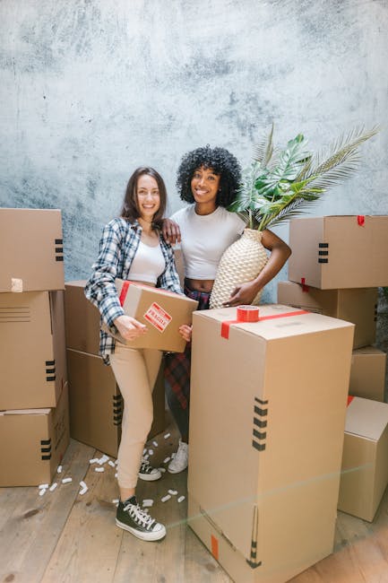 Two women are standing indoors among several large packed cardboard moving boxes, some sealed with red and black packing tape. One woman, with shoulder-length brown hair and wearing a plaid shirt over a white top and beige pants, holds a small box with a red label. The other woman, with curly black hair and wearing a white top and dark plaid skirt, is holding a large plastic-wrapped leafy plant in a woven basket. The boxes are stacked against a textured, light grey wall, and the wooden floor is partially covered with packing material. The scene depicts a home relocation process with careful packing and furniture transport preparation, as [COMPANY_NAME] assists with house removals or moving services, ensuring efficient furniture transport and packing during the move.