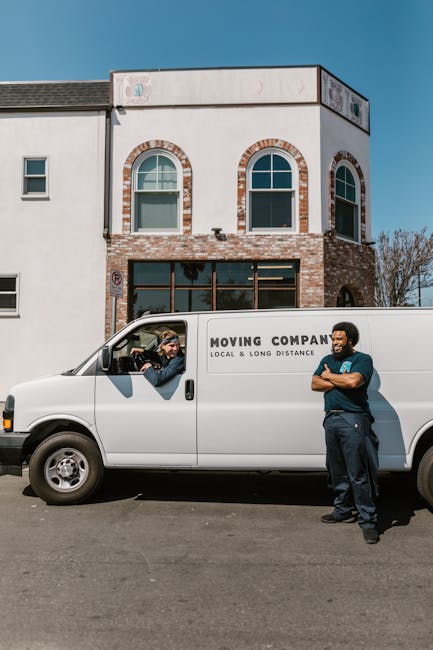 A white moving van parked on the street outside a two-storey residential building with a brick and white exterior under a clear blue sky. The van has the logo and text indicating it belongs to a local moving company, with the phrase 'MOVING COMPANY LOCAL & LONG DISTANCE' visible on the side. Inside the driver’s cabin, a person wearing a cap and sunglasses is seen handing over or receiving documents from a person standing on the pavement next to the van. The person on the pavement, dressed in dark clothing with arms crossed, appears to be a mover or company staff member, engaging in a conversation with the driver. Behind the parked van, the building features three windows on the upper floor; the central window has an arched top, while the lower level appears to have large glass doors or windows, likely leading to a commercial or residential space. The scene captures a moment of home relocation preparation or coordination, consistent with furniture transport and packing and moving activities by [COMPANY_NAME], as part of house removals services.
