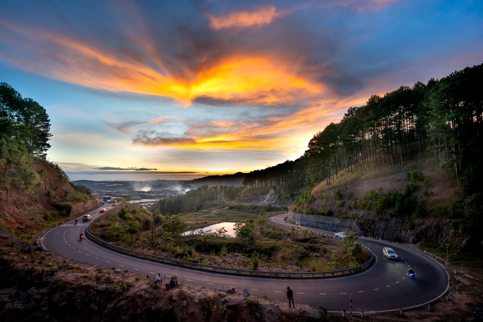 A winding mountain road with multiple sharp bends, situated between steep, wooded hillsides covered in dense green trees, leading towards a distant horizon. On the road, several vehicles including cars and vans are visible, some approaching the curves while others are positioned along the roadside. To the left side of the road, a small group of people are sitting on the rocky terrain, possibly observing the scenery. The sky overhead features vibrant orange, yellow, and pink hues from a setting or rising sun, with scattered clouds creating a dramatic atmosphere. The scene suggests a remote, scenic area suitable for travel and transportation, which aligns with themes of home relocation and furniture transport services, such as those offered by Man with Van Forest Hill.