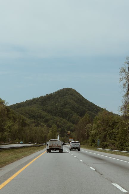 The image shows a view of a two-lane road during daylight, with a white van and a white car traveling side by side towards a green, forested hill in the background. The road is bordered by metal guardrails and trees on both sides, and the sky is partly cloudy. This scene captures the journey aspect of house removals or furniture transport, with vehicles actively moving along a scenic route toward a residential or urban area, aligning with services offered by Man with Van Forest Hill. The road surface appears to be in good condition, and the vehicles are positioned in the center lanes, suggesting a typical loading process or transportation phase during a home relocation.