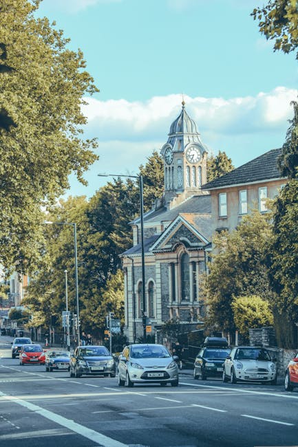 A row of parked cars on a busy street in Forest Hill with a church featuring a prominent clock tower and a domed roof visible in the background. The street is lined with mature trees providing shade, and the sky is bright with scattered clouds. The scene is set during daytime with natural lighting. In the foreground, the cars are parked along the curb, while some vehicles are driving along the road. The surrounding buildings are residential with brick facades, and there are streetlights and traffic signs nearby. This image visually represents a typical urban environment where house removals or furniture transport could take place, emphasizing the context of home relocation logistics in Forest Hill, as handled by Man with Van Forest Hill.